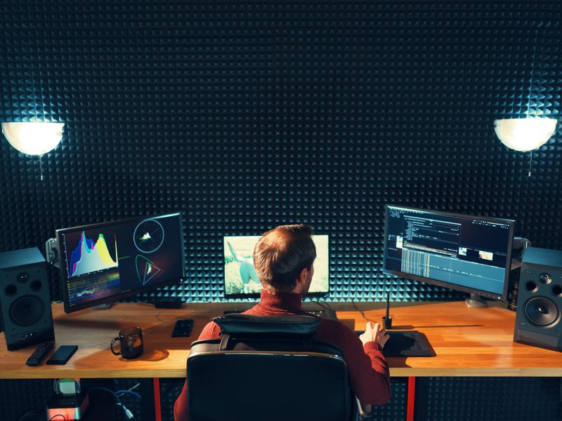 Video editing. Professional editor adding special sound effects. Back view of young man watching graphs on monitors. Copy space on gray wall